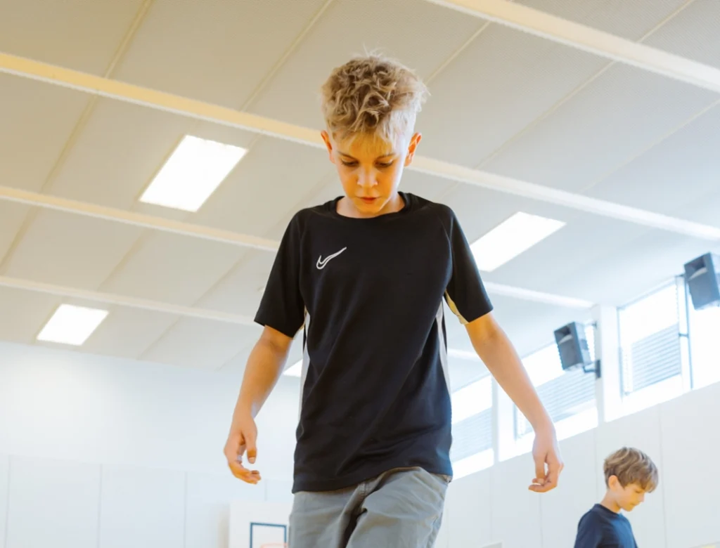 Junge geht konzentriert und mit gesenktem Blick durch eine Turnhalle, im Hintergrund ein weiteres Kind